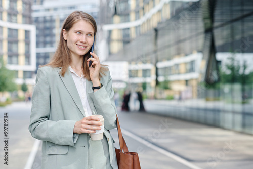 Cheerful young Caucasian woman wearing stylish formal suit holding cup of coffee and talking on phone on way to work