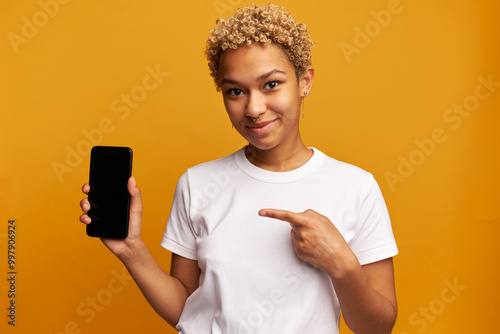 Young black girl in white holding smartphone in hands, showing you black copy space display for your advertising content, promoting and recommending new mobile product or app, isolated on yellow
