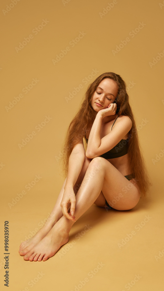Portrait of tender, elegant young woman with long hair, sitting on floor in underwear and posing against sandy color studio background. Concept of natural female beauty, body and skin care, health Portrait of tender, elegant young woman with long hair, sitting on floor in underwear and posing against sandy color studio background. Concept of natural female beauty, body and skin care, health