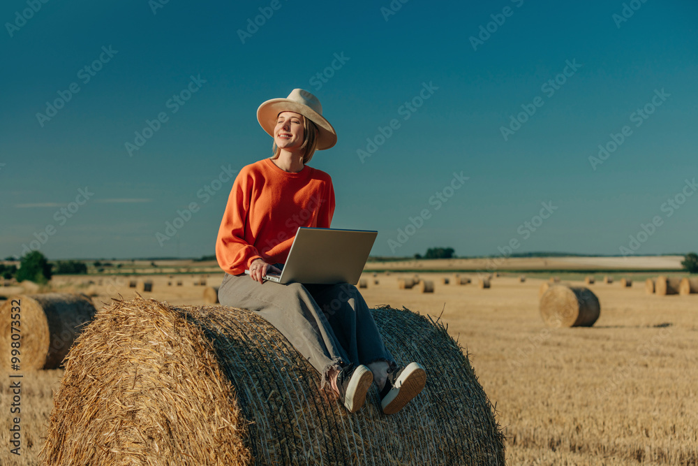 Happy woman wearing hat and sitting with laptop on hay bale in field Happy woman wearing hat and sitting with laptop on hay bale in field