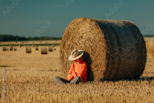 Sad woman wearing hat sitting near hay bale on field