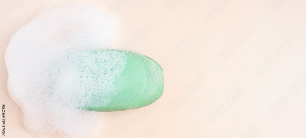 Fototapeta premium Top view of single antibacterial hygienic organic soap bar of green colour wet soapy and covered with foam bubbles laying on beige background with copy space used as toiletries at bath for skin care