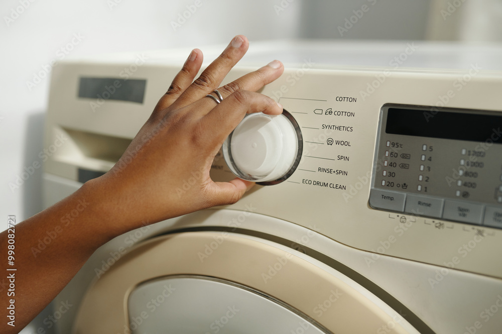 Close-up view of person adjusting settings on a front-loading washing machine. Display showing various fabric options with hand turning dial Close-up view of person adjusting settings on a front-loading washing machine. Display showing various fabric options with hand turning dial