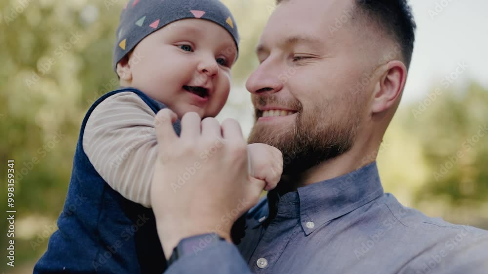 A loving father smiles as he holds his adorable baby boy. The child, in a colorful hat, shows a big, happy smile in a sunlit outdoor setting. Joyful Father Holding Cute Baby Boy in Natural Setting A loving father smiles as he holds his adorable baby boy. The child, in a colorful hat, shows a big, happy smile in a sunlit outdoor setting. Joyful Father Holding Cute Baby Boy in Natural Setting