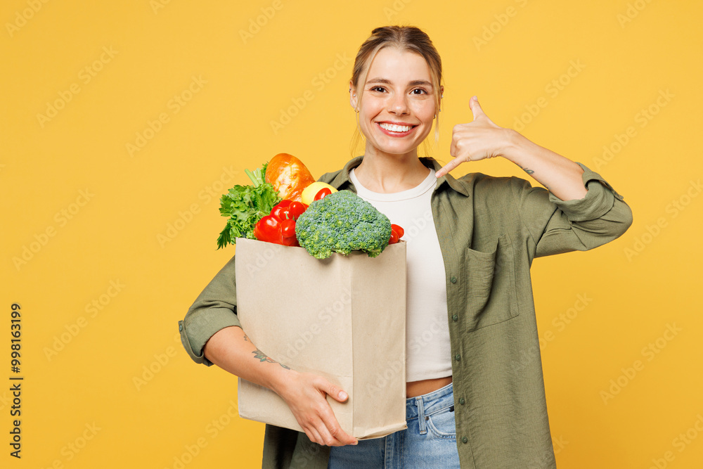 Young woman wear casual clothes hold bag for takeaway mock up with food products do phone gesture like says call me back isolated on plain yellow background. Delivery service from shop or restaurant. Young woman wear casual clothes hold bag for takeaway mock up with food products do phone gesture like says call me back isolated on plain yellow background. Delivery service from shop or restaurant.