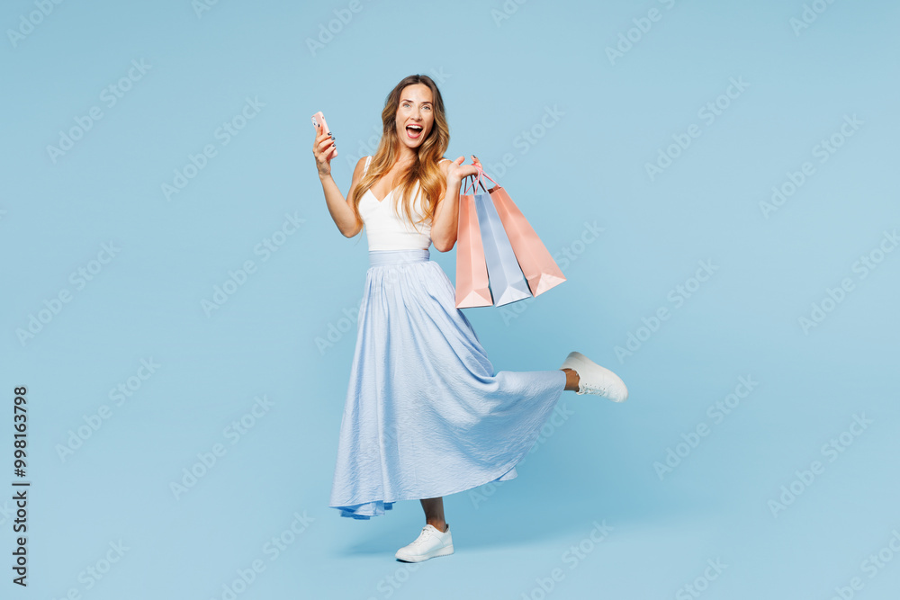 Full body young surprised fun woman wear casual clothes hold in hand paper package bags after shopping use mobile cell phone isolated on plain light blue background. Black Friday sale buy day concept. Full body young surprised fun woman wear casual clothes hold in hand paper package bags after shopping use mobile cell phone isolated on plain light blue background. Black Friday sale buy day concept.