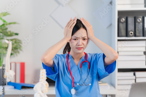 Exhaustion in the Medical Field: A woman doctor in blue scrubs with a stethoscope, holds her head with both hands, expressing the exhaustion and stress of her profession. photo