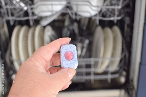 A woman holds a capsule of detergent against the backdrop of a dishwasher with dirty dishes. photo