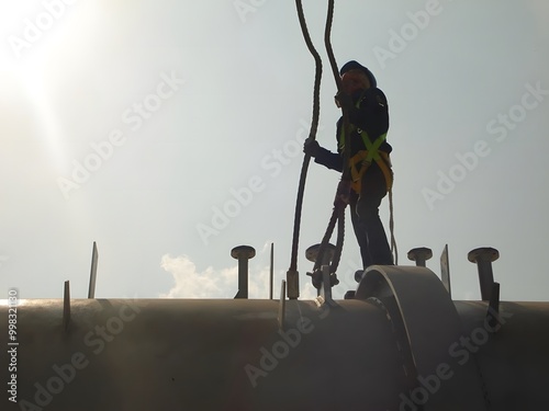 A silhouetted worker in a safety harness and helmet is perched on a large pipe, performing maintenance. The bright sky highlights the height and the critical importance of safety measures.