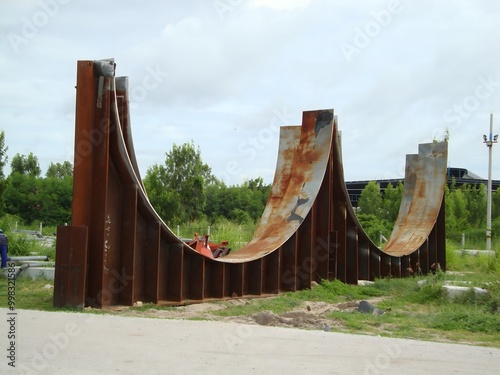 Three large, rusty, curved metal structures stand upright in a field, resembling giant half-pipes. Construction equipment is visible in the background, hinting at an unfinished industrial project 