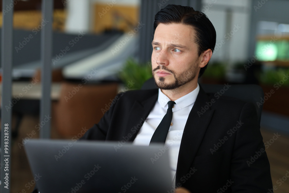 A man in a suit and tie is sitting at a desk with a laptop open in front of him. He is focused on his work, possibly preparing for a meeting or working on a project A man in a suit and tie is sitting at a desk with a laptop open in front of him. He is focused on his work, possibly preparing for a meeting or working on a project