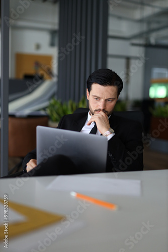 A man in a suit is sitting at a desk with a laptop open in front of him. He is deep in thought, possibly working on a project or problem. Concept of focus and concentration