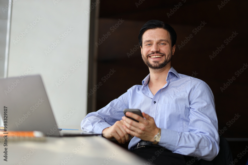 A man is sitting at a desk with a laptop and a cell phone. He is smiling and he is happy A man is sitting at a desk with a laptop and a cell phone. He is smiling and he is happy
