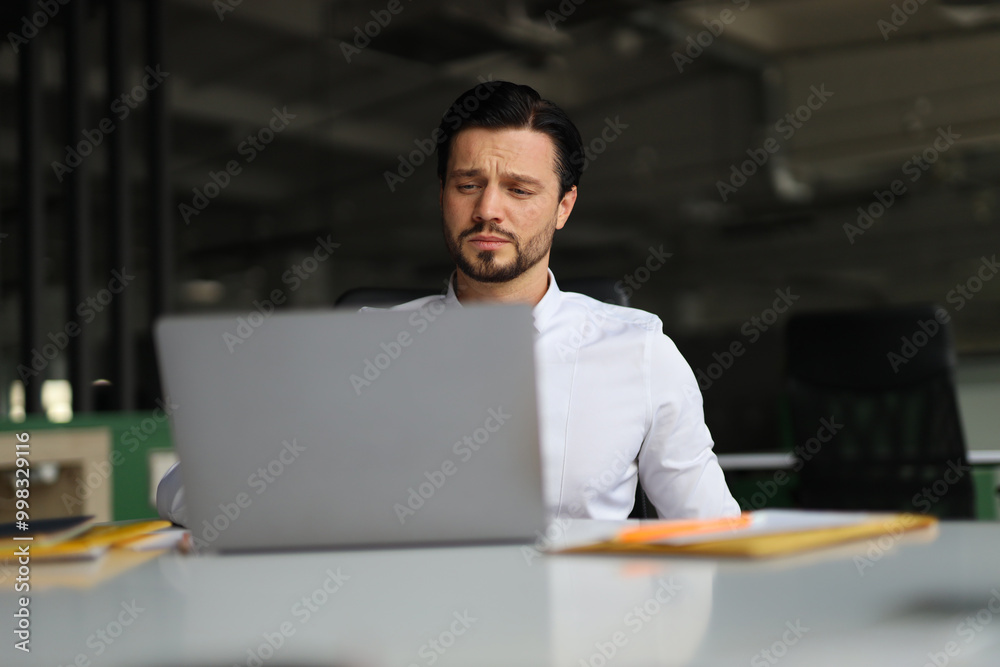 A man is sitting at a desk with a laptop open in front of him. He is wearing a white shirt and he is focused on his work. Concept of productivity and concentration A man is sitting at a desk with a laptop open in front of him. He is wearing a white shirt and he is focused on his work. Concept of productivity and concentration