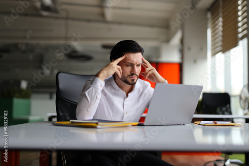 A man is sitting at a desk with a laptop and a stack of papers. He is looking at the laptop with a furrowed brow, possibly indicating that he is stressed or overwhelmed