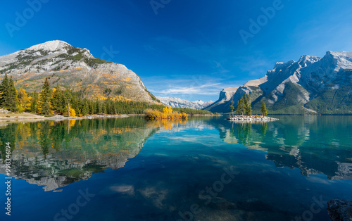 Banff National Park Lake Minnewanka autumn landscape. Alberta, Canada. Majestic mountains, forests in yellow green color against blue sky reflected on the water. Beautiful natural scenery.