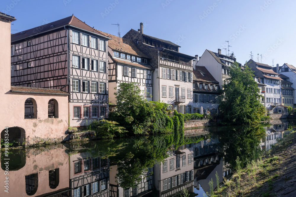 Naklejka premium Strasbourg, France - August 28th 2024 : View of the Tanner's Quarter with half-timbered buildings and the Ill river and reflections on the water.