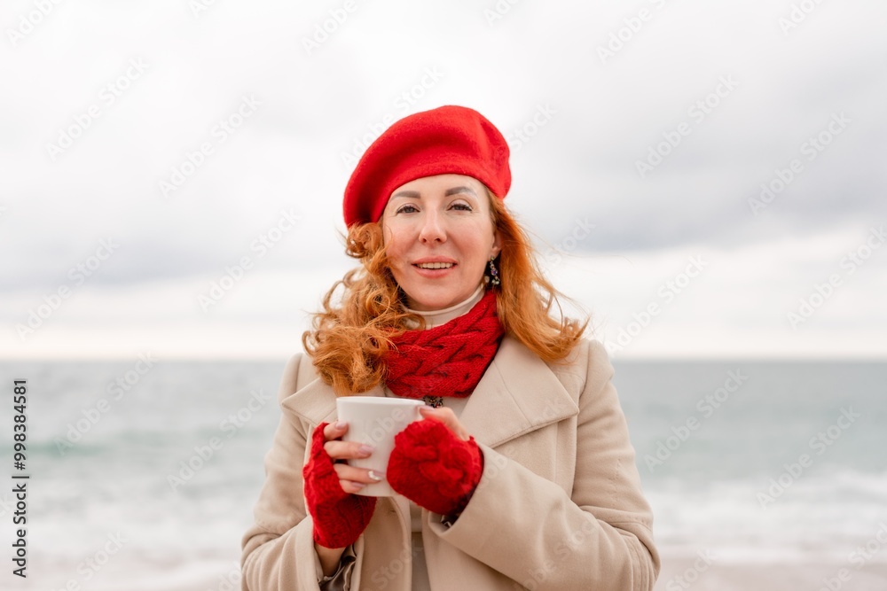 Obraz premium Woman beach in red beret scarf and mitts holding a cup of tea in his hands. Depicting beach relaxation and cozy attire. Walks by the sea