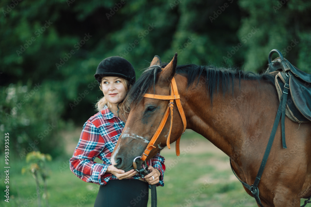 Obraz premium Happy blonde with horse in forest. Woman and a horse walking through the field during the day. Dressed in a plaid shirt and black leggings.