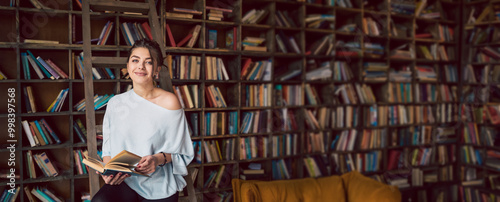 Beautiful young smiling woman holding book and looking at the camera in a home library with many bookshelves in the background. Student exam preparation. Long horizontal banner 