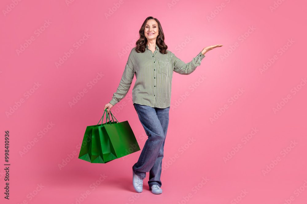 Fototapeta premium Full length photo of charming positive woman wear striped shirt holding shoppers holding arm empty space isolated pink color background