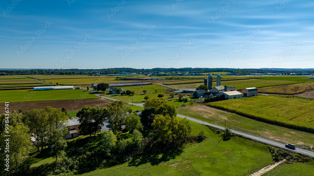 Expansive farmland stretches under a bright blue sky, featuring neatly arranged fields and silos near a rural road. The landscape showcases vibrant greenery and agricultural structures. Expansive farmland stretches under a bright blue sky, featuring neatly arranged fields and silos near a rural road. The landscape showcases vibrant greenery and agricultural structures.