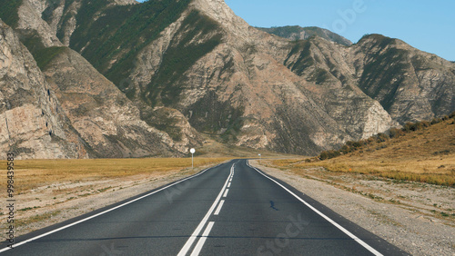 Driving on empty highway of black asphalt and white road markings at sunny day, road going into distance through desert prairie at fall to rocky mountain massive on horizon in wild. Pov moving car