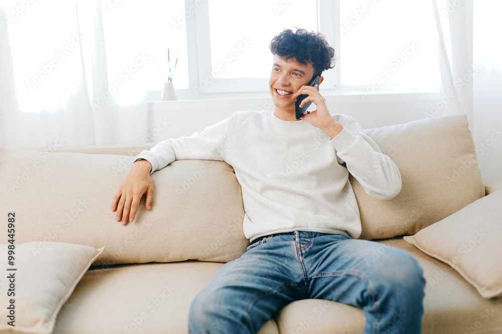 smiling young man talking on smartphone, casual atmosphere, sitting on a light couch, indoor setting, natural light, comfortable and relaxed ambiance smiling young man talking on smartphone, casual atmosphere, sitting on a light couch, indoor setting, natural light, comfortable and relaxed ambiance