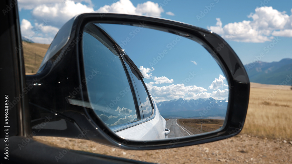 Fototapeta premium Car side rearview mirror closeup. View mountain range with snow capped summits from driving car vehicle on highway road passing through valley of foothills on sunny day. Road trip in summer holidays