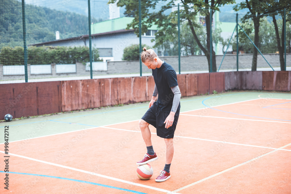 Young man with a man bun juggles a football on a vibrant orange court during a sunny day. Wearing casual sportswear, he focuses on controlling the ball with precision in a natural setting Young man with a man bun juggles a football on a vibrant orange court during a sunny day. Wearing casual sportswear, he focuses on controlling the ball with precision in a natural setting
