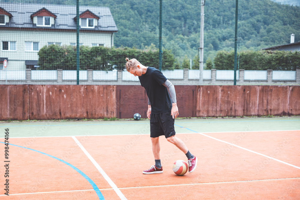 Obraz premium Young man with a man bun juggles a football on a vibrant orange court during a sunny day. Wearing casual sportswear, he focuses on controlling the ball with precision in a natural setting