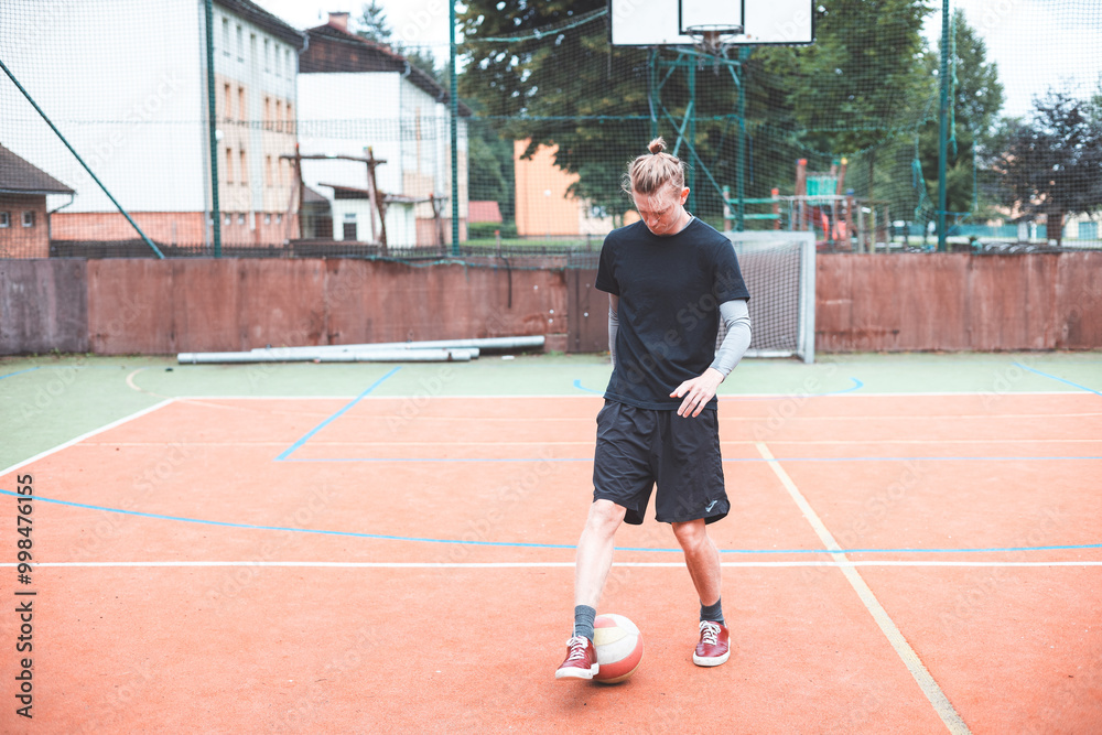 Fototapeta premium Young man with a man bun juggles a football on a vibrant orange court during a sunny day. Wearing casual sportswear, he focuses on controlling the ball with precision in a natural setting
