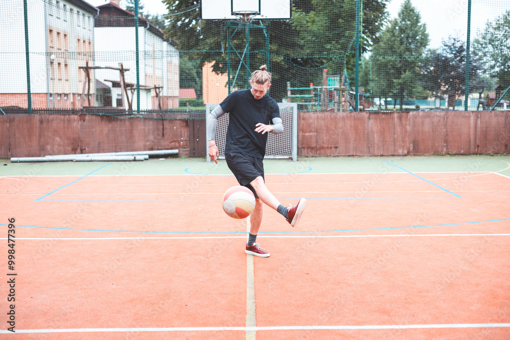Obraz premium Young man with a man bun juggles a football on a vibrant orange court during a sunny day. Wearing casual sportswear, he focuses on controlling the ball with precision in a natural setting