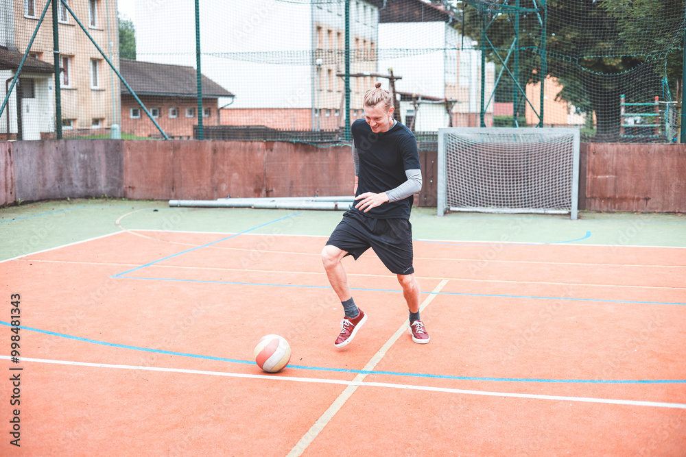 Obraz premium Young man focuses on controlling a football on an outdoor court. He carefully juggles the ball with precision, surrounded by an urban backdrop of buildings and a goal, combining sports and skill