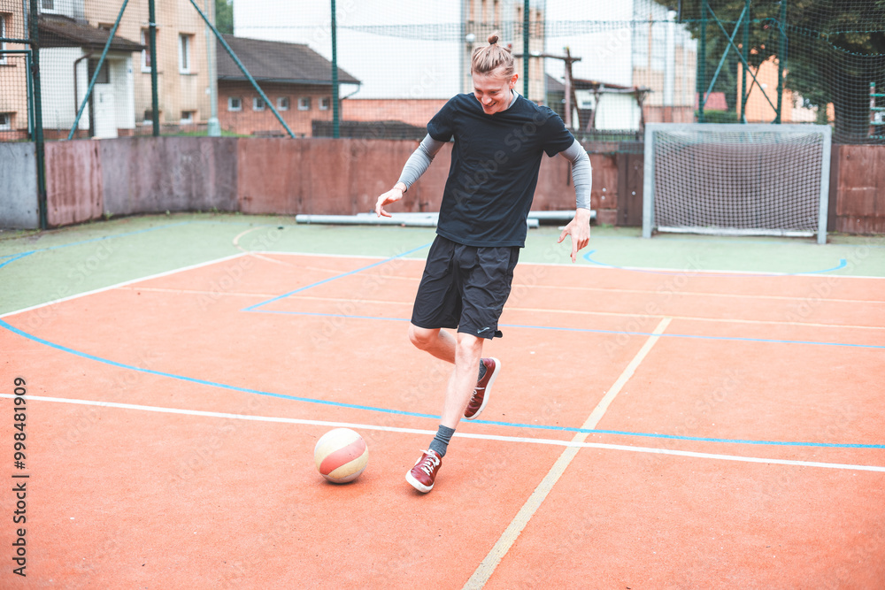 Obraz premium Young man focuses on controlling a football on an outdoor court. He carefully juggles the ball with precision, surrounded by an urban backdrop of buildings and a goal, combining sports and skill