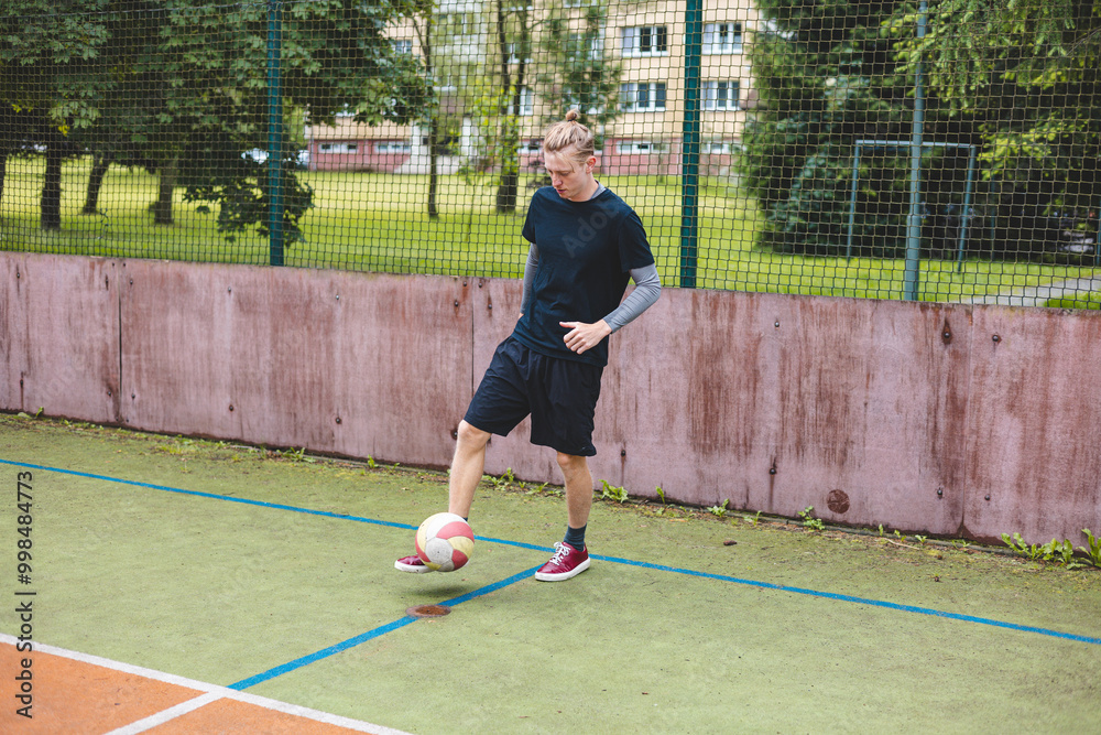 Young man focuses on controlling a football on an outdoor court. He carefully juggles the ball with precision, surrounded by an urban backdrop of buildings and a goal, combining sports and skill Young man focuses on controlling a football on an outdoor court. He carefully juggles the ball with precision, surrounded by an urban backdrop of buildings and a goal, combining sports and skill