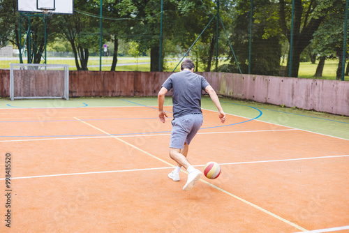 Young man dribbles a football with precision on an outdoor court, practicing his footwork in a casual environment. The court is surrounded by nature, providing a peaceful backdrop