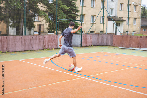 Young man takes a powerful shot on goal while playing football on an outdoor court. Wearing casual attire, he shows intensity and focus in a dynamic moment with an urban backdrop behind him