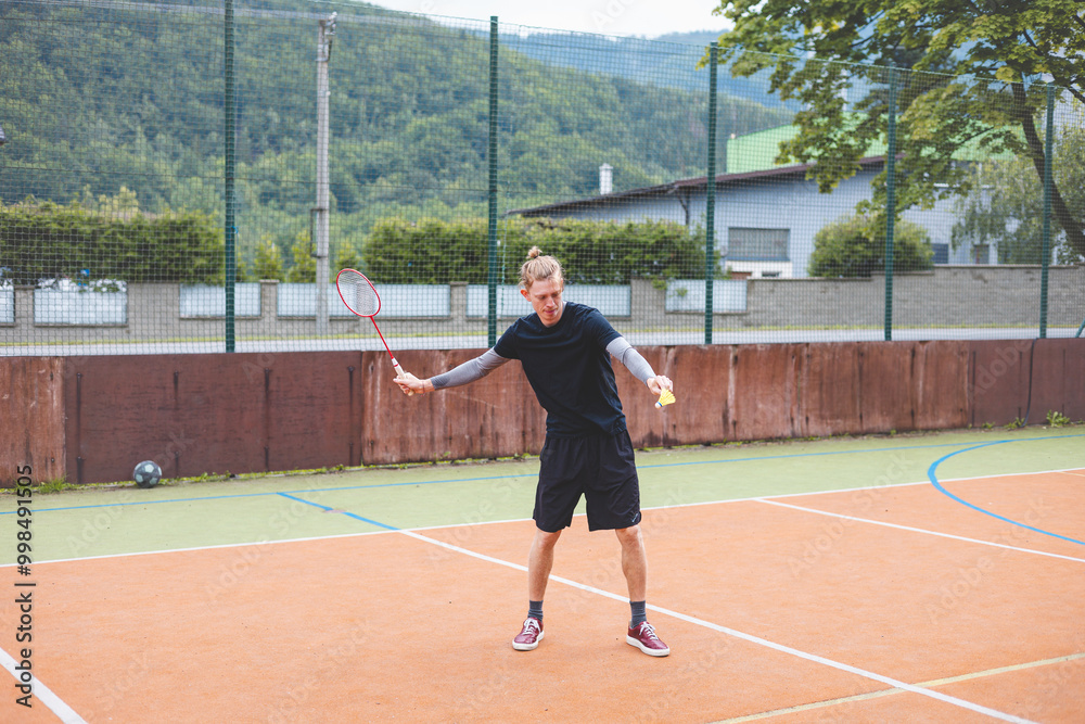 Badminton player stands ready to serve on an outdoor court, holding a racket and shuttlecock. Dressed in casual sportswear, he focuses on the game amid a scenic, natural setting Badminton player stands ready to serve on an outdoor court, holding a racket and shuttlecock. Dressed in casual sportswear, he focuses on the game amid a scenic, natural setting