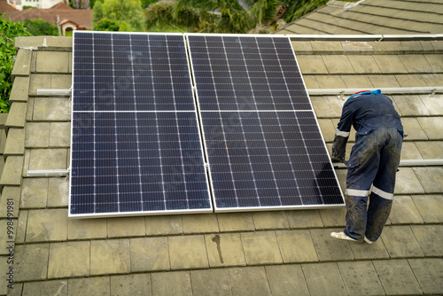 Builder mounting photovoltaic solar modules on roof of house. Back view of man engineer in helmet installing solar panel system outdoors. Concept of alternative and renewable energy.