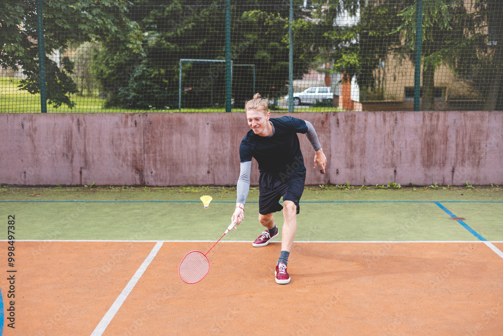 Badminton player strikes the shuttlecock during a game on an outdoor court, showcasing precise technique and athletic movement. The player is focused, surrounded by trees and an urban backdrop Badminton player strikes the shuttlecock during a game on an outdoor court, showcasing precise technique and athletic movement. The player is focused, surrounded by trees and an urban backdrop