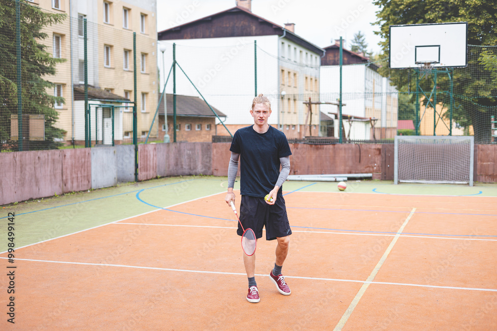 Obraz premium Badminton player picks up a shuttlecock during a game on an outdoor court. The casual moment takes place in an urban setting, with surrounding buildings and a basketball hoop in the background