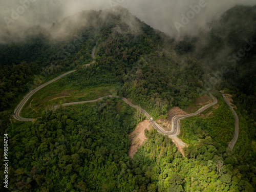 A rural road winds through a serene mountain vista, bordered by lush green forest under a blue sky at dawn in Doi Phuka National Park, Nan Province, Thailand photo