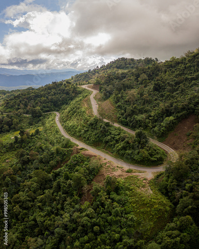 A rural road winds through a serene mountain vista, bordered by lush green forest under a blue sky at dawn in Doi Phuka National Park, Nan Province, Thailand photo