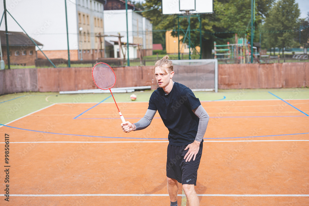 Obraz premium Badminton player strikes the shuttlecock during a game on an outdoor court, showcasing precise technique and athletic movement. The player is focused, surrounded by trees and an urban backdrop