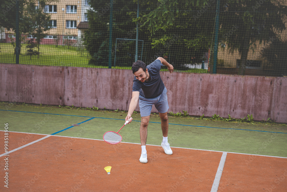 Badminton player stretches to retrieve a low shuttlecock during an intense game on an outdoor court. His athletic stance shows focus and determination, with an urban setting in the background Badminton player stretches to retrieve a low shuttlecock during an intense game on an outdoor court. His athletic stance shows focus and determination, with an urban setting in the background