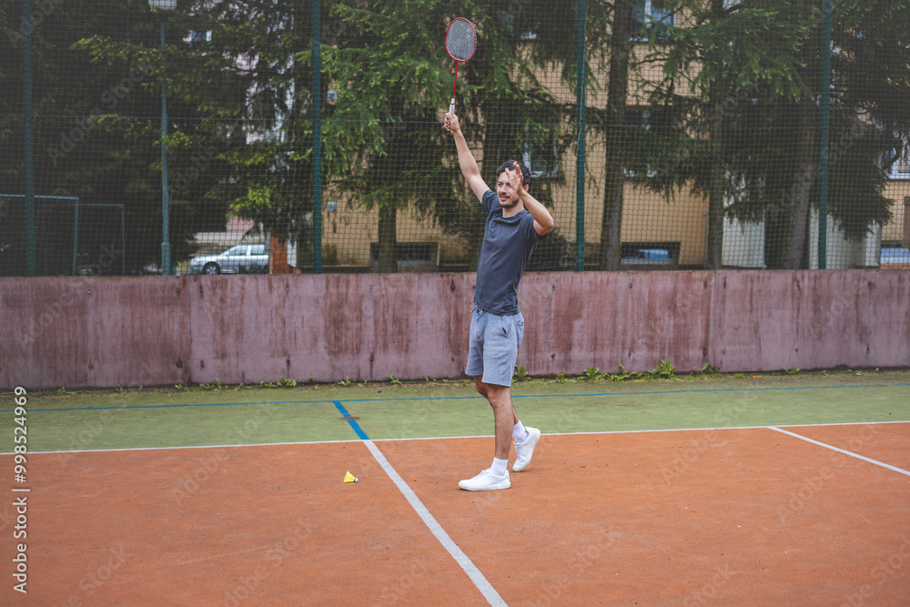 Badminton player strikes the shuttlecock during a game on an outdoor court, showcasing precise technique and athletic movement. The player is focused, surrounded by trees and an urban backdrop Badminton player strikes the shuttlecock during a game on an outdoor court, showcasing precise technique and athletic movement. The player is focused, surrounded by trees and an urban backdrop