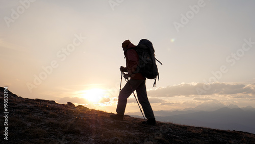 Man climber with backpack and trekking sticks poles raises up to top of cliff mountain. Walking at sunrise or sunset, sun rays shine background. Dressed in warm trekking clothes hiking. Shot in motion