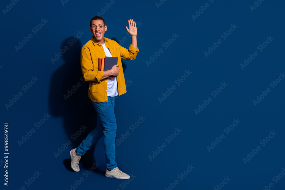Photo of young smart guy wearing eyeglasses holding books preparing to exam waving hand welcome isolated on dark blue color background Photo of young smart guy wearing eyeglasses holding books preparing to exam waving hand welcome isolated on dark blue color background