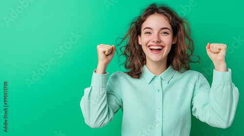 A woman with a triumphant grin, fists raised in victory, standing against a pastel green background, celebrating her success.